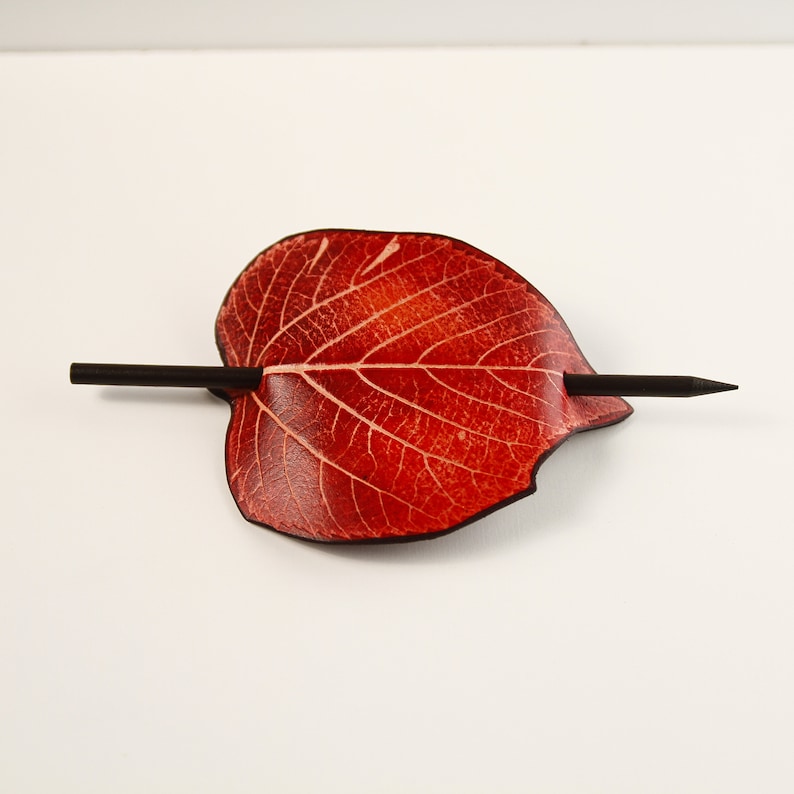 May include: A red leaf-shaped hair barrette with a dark wooden stick. The barrette has a detailed vein pattern and a smooth, polished finish. The hair accessory is designed to hold hair in place.