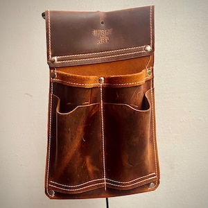 May include: A brown leather tool belt with multiple pockets and white stitching. The top flap is embossed with the text "CURLY ART". The belt has a rustic, worn appearance and is designed to be worn around the waist.