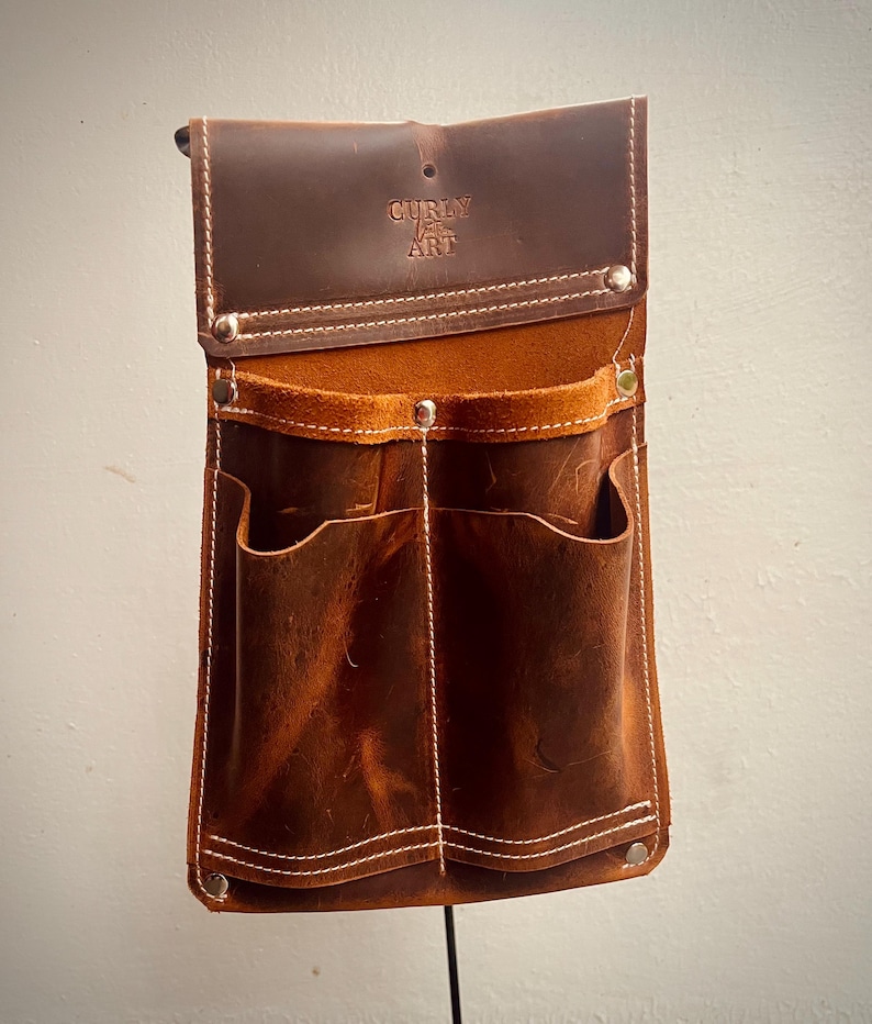 May include: A brown leather tool belt with multiple pockets and white stitching. The top flap is embossed with the text "CURLY ART". The belt has a rustic, worn appearance and is designed to be worn around the waist.