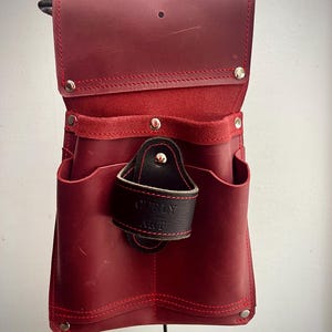 May include: A red leather tool belt with multiple pockets and a black leather loop. The belt features red stitching and silver rivets. The text "CURLY ART" is visible on the black loop.