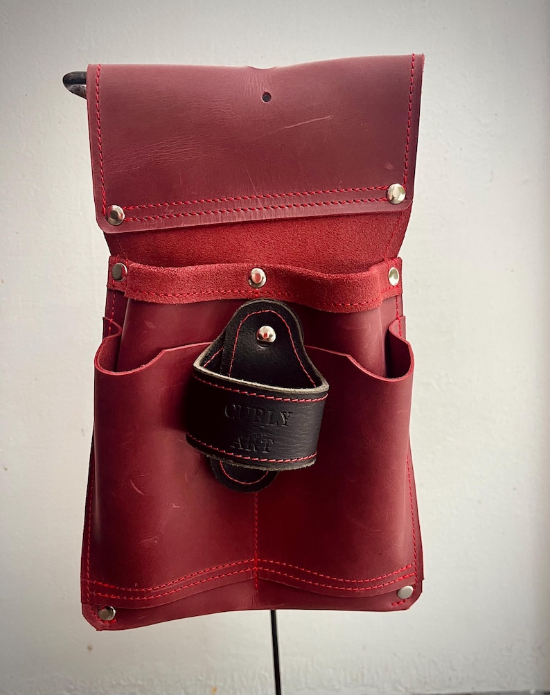 May include: A red leather tool belt with multiple pockets and a black leather loop. The belt features red stitching and silver rivets. The text "CURLY ART" is visible on the black loop.