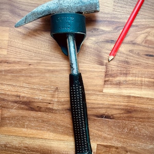 May include: A claw hammer with a black textured handle and a silver head, next to a red pencil. The hammer has the text "CURLY ART" on the head. The tools are on a wooden surface.