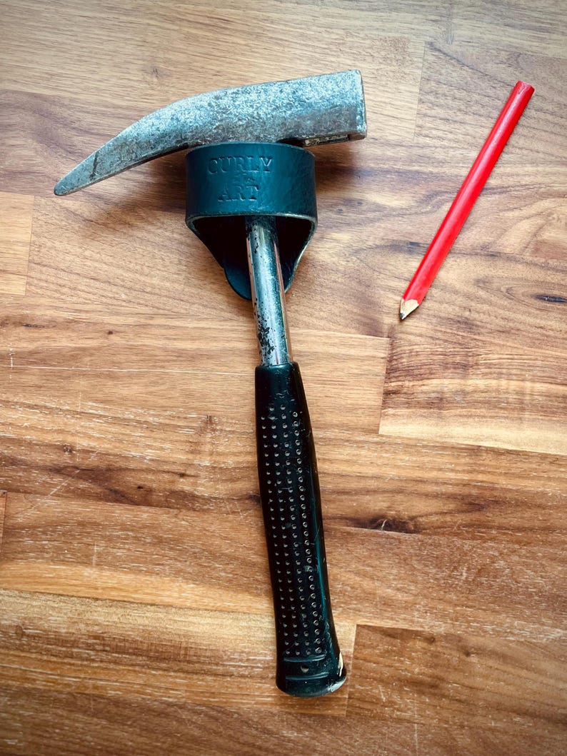 May include: A claw hammer with a black textured handle and a silver head, next to a red pencil. The hammer has the text "CURLY ART" on the head. The tools are on a wooden surface.