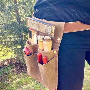 May include: A brown leather tool belt with multiple pockets, holding various carpentry tools. The belt is worn around the waist and features a brown leather strap. Tools include a level, ruler, pencils, and screwdrivers.