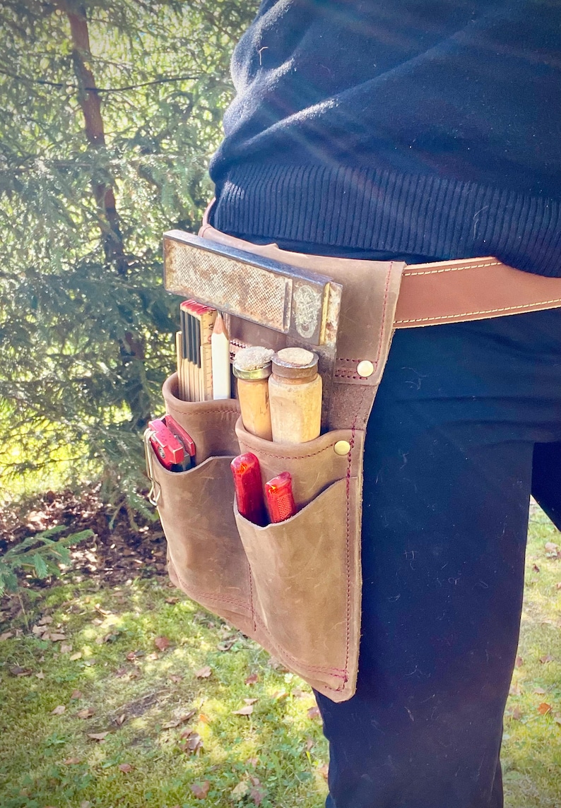 May include: A brown leather tool belt with multiple pockets, holding various carpentry tools. The belt is worn around the waist and features a brown leather strap. Tools include a level, ruler, pencils, and screwdrivers.
