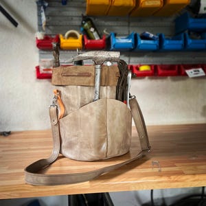 Può includere: Una borsa porta attrezzi in pelle color cuoio con tracolla, piena di attrezzi tra cui un martello e delle pinze. La borsa ha più tasche e una finitura invecchiata, che suggerisce uno stile vintage o rustico. Lo sfondo mostra un'officina.