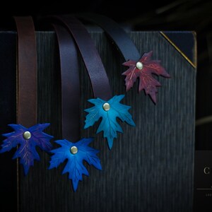 Puede incluir: Cuatro marcapáginas de cuero con diseños de hojas de arce. Las hojas son en tonos de azul, morado y rojo. Los marcapáginas están sobre una superficie de madera oscura. El texto "Curly Leather Art" es visible en la esquina inferior derecha.
