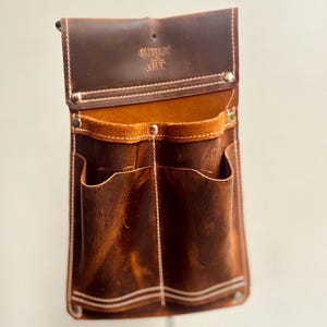 May include: A brown leather tool belt with multiple pockets and the text "Curly Art" embossed on the flap. The belt has white stitching and metal rivets. The interior is a lighter brown color.