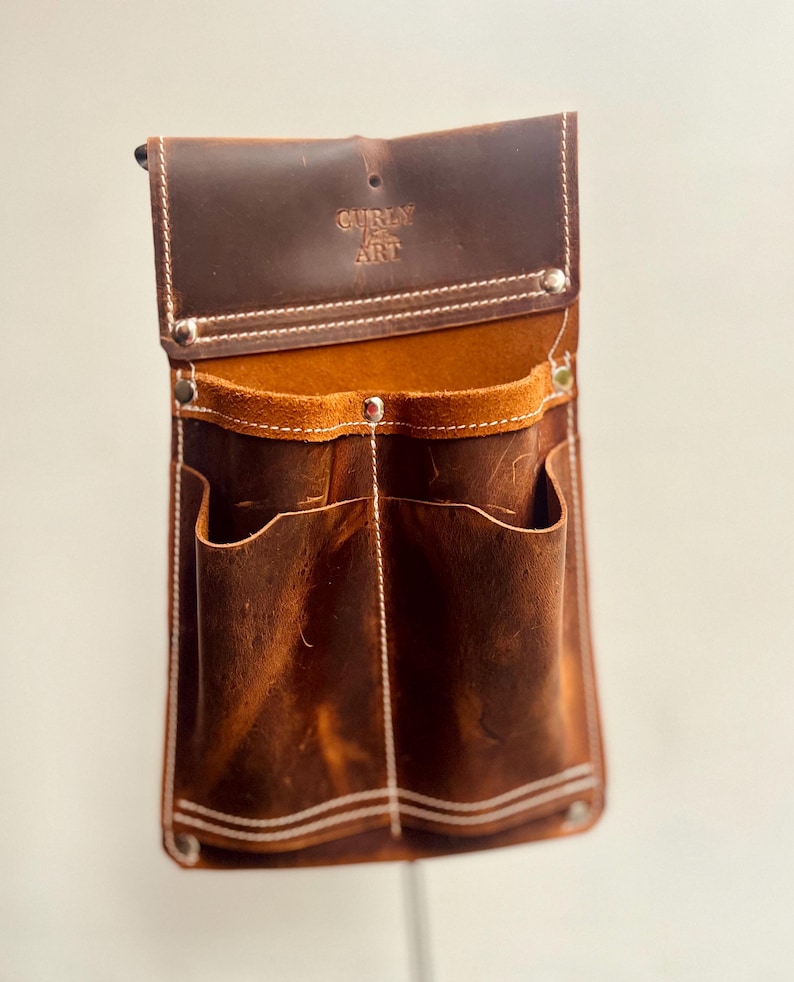 May include: A brown leather tool belt with multiple pockets and the text "Curly Art" embossed on the flap. The belt has white stitching and metal rivets. The interior is a lighter brown color.
