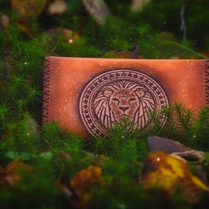 Könnte beinhalten: Eine braune Ledergeldbörse mit einem geprägten Löwenmotiv auf der Vorderseite. Die Geldbörse liegt auf einem Bett aus grünem Moos und Blättern.