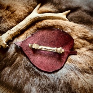 May include: A burgundy leaf-shaped hair clip with a gold-colored barrette clasp. The clip is set on a backdrop of brown and tan fur, with a section of a deer antler in the background. The overall aesthetic is rustic and natural.