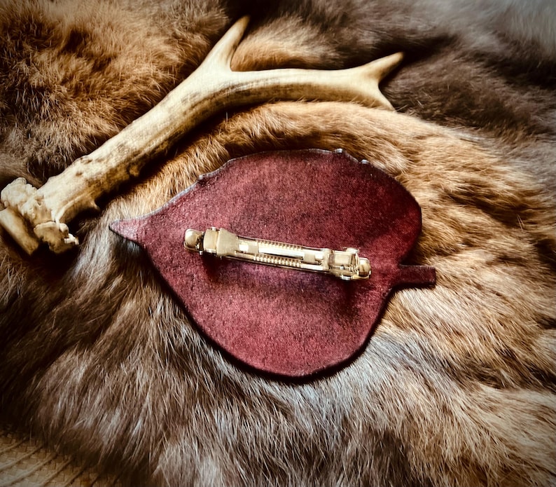 May include: A burgundy leaf-shaped hair clip with a gold-colored barrette clasp. The clip is set on a backdrop of brown and tan fur, with a section of a deer antler in the background. The overall aesthetic is rustic and natural.