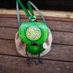 May include: A handmade leather backpack with a green flap and a beige body. The green flap features a gold Celtic knot design. The backpack has green straps and a drawstring closure. The bag is sitting on a wooden surface.
