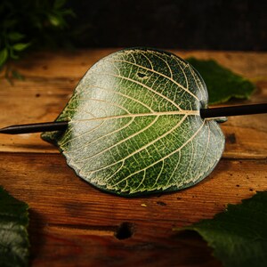 May include: A handcrafted hair accessory featuring a green leaf design with a dark brown stick. The leaf has detailed veining and a natural appearance. The hair accessory is set against a wooden background.