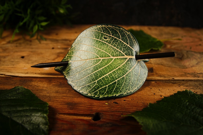 May include: A handcrafted hair accessory featuring a green leaf design with a dark brown stick. The leaf has detailed veining and a natural appearance. The hair accessory is set against a wooden background.