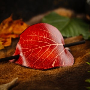 May include: A handcrafted hair barrette shaped like a red leaf with a dark wooden stick. The leaf has detailed veining and is set against a backdrop of autumn leaves and greenery. A unique hair accessory.