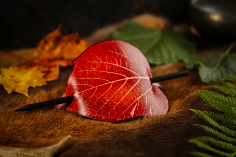 May include: A handcrafted hair barrette shaped like a red leaf with a dark wooden stick. The leaf has detailed veining and is set against a backdrop of autumn leaves and greenery. A unique hair accessory.