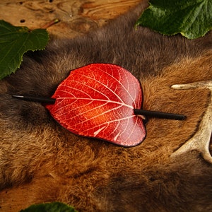 May include: A red leaf-shaped hair barrette with a black stick pin, resting on a brown fur surface. The leaf has white vein detailing. Green leaves and a deer antler are also visible.