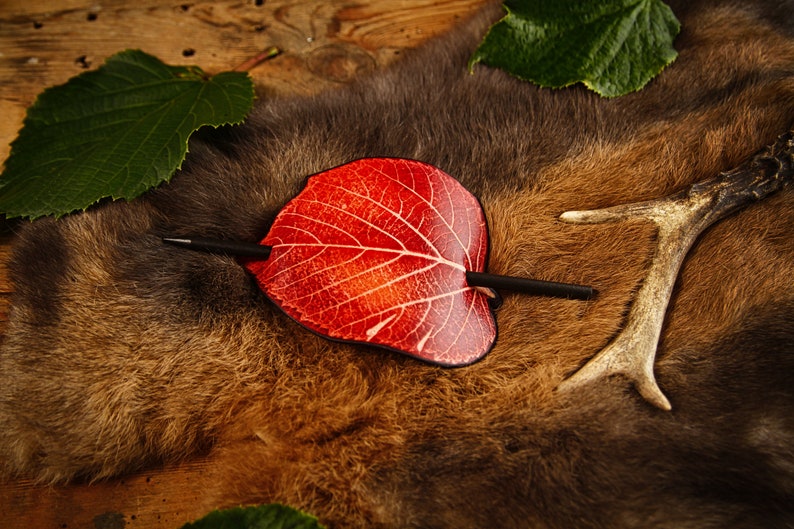 May include: A red leaf-shaped hair barrette with a black stick pin, resting on a brown fur surface. The leaf has white vein detailing. Green leaves and a deer antler are also visible.