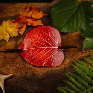 May include: A red leaf-shaped hair barrette with a black stick pin, resting on a brown fur-like surface. The barrette is surrounded by autumn leaves and greenery, creating a natural, rustic aesthetic.