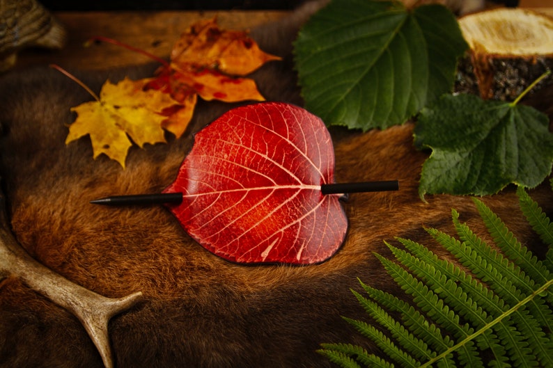 May include: A red leaf-shaped hair barrette with a black stick pin, resting on a brown fur-like surface. The barrette is surrounded by autumn leaves and greenery, creating a natural, rustic aesthetic.