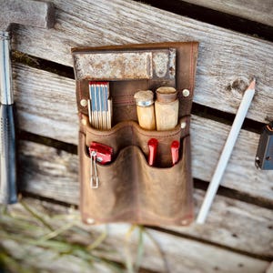 May include: A brown leather tool belt with multiple pockets, holding a folding ruler, matches, small wooden containers, and red tools. A white pencil and a hammer are visible. The belt is on a wooden surface.