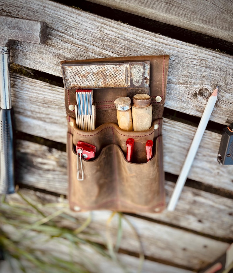 May include: A brown leather tool belt with multiple pockets, holding a folding ruler, matches, small wooden containers, and red tools. A white pencil and a hammer are visible. The belt is on a wooden surface.