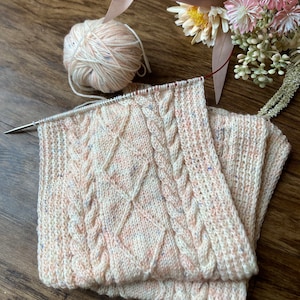 May include: A close-up of a knitted fabric with a cable stitch pattern. The fabric is a light pink and white colour with a speckled texture. A ball of yarn is visible in the background.