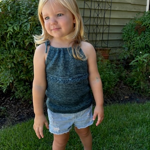 May include: A young child wearing a teal blue knitted halter top and light blue denim shorts. The child is standing on a green grassy lawn.