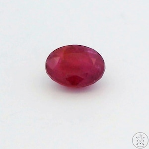 May include: An oval-shaped, faceted ruby gemstone. The ruby is a deep, saturated red color and is set against a white background. The gemstone is a polished, precious stone.