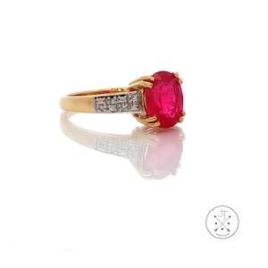 May include: A gold ring featuring a large, oval-cut ruby gemstone. The ring has a band with small, clear accent stones on each side of the ruby. The ring is displayed on a reflective white surface.