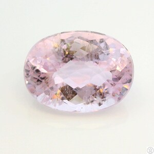 May include: A light pink, oval-cut gemstone with internal facets. The gem has a soft, translucent appearance with subtle variations in color. The overall shape is symmetrical, and the surface reflects light, creating a sparkling effect. The gemstone is set against a white background.