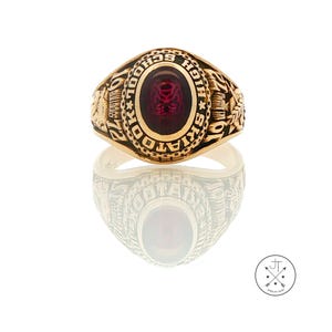 May include: A gold-toned class ring featuring a central oval-shaped deep red stone with an embossed bulldog emblem. The bezel is engraved with the text SKIATOOK HIGH SCHOOL and the year 1972, with intricate relief details on the side shanks.