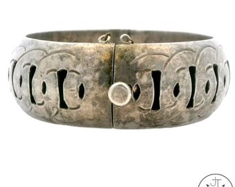 May include: A silver-toned hinged bangle bracelet. The bracelet features a series of cut-out shapes and a decorative pattern. The clasp is a small, round button. The bracelet is a wide cuff style.
