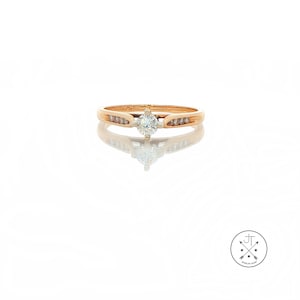 May include: A gold ring with a central diamond and smaller diamonds on the band. The ring is set against a white background, with a reflection visible below. The ring is a classic design, suitable for engagement or everyday wear.