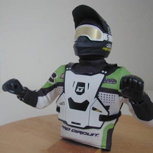Peut inclure: Une figurine de motocross noire et verte portant un plastron blanc avec le texte "Pro Circuit". La figurine porte un casque noir avec une visière dorée et une combinaison de course noire et blanche.