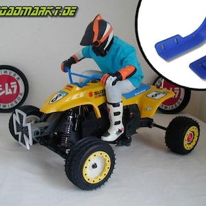 Könnte beinhalten: Ein gelbes Suzuki-Spielzeug-Quad mit einer Fahrerfigur, die einen blau-orangenen Helm trägt. Das Quad hat schwarze Räder mit gelben Felgen und eine schwarz-weiße kreuzförmige Stoßstange. Das Spielzeug befindet sich auf einer weißen Oberfläche mit einem rot-schwarzen Yoshimura-Logo im Hintergrund.