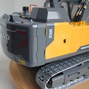 Op de afbeelding: Gedetailleerd model van een Volvo EC160E graafmachine. Het model heeft een grijs-geel kleurenschema, met het Volvo-logo prominent aanwezig. De graafmachine heeft een gedetailleerd rupsbandsysteem en een realistisch ontwerp.