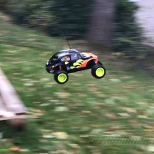 May include: A black and orange remote control car with yellow wheels is in mid-air, flying over a wooden pallet. The car has a flame design on the side.