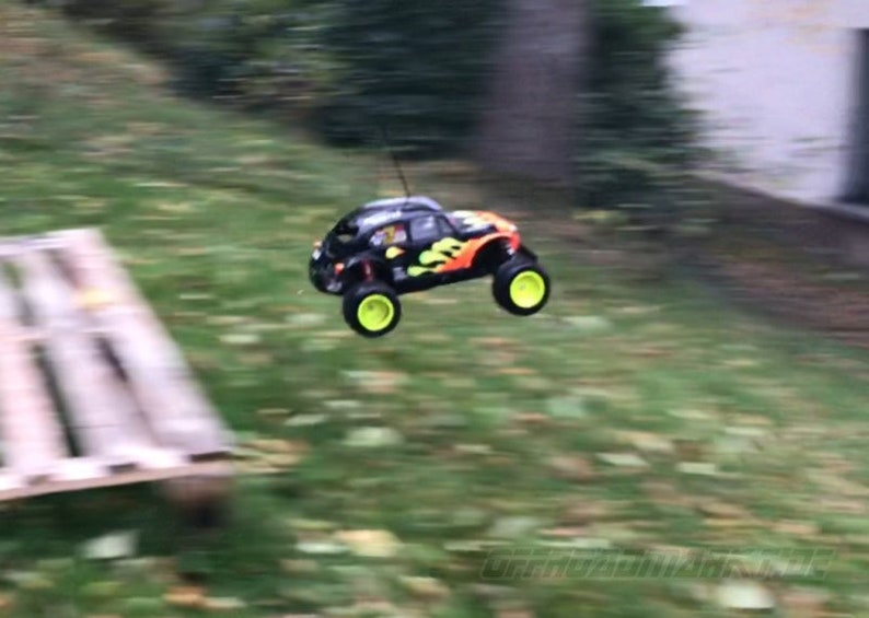 May include: A black and orange remote control car with yellow wheels is in mid-air, flying over a wooden pallet. The car has a flame design on the side.