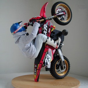 Könnte beinhalten: Ein rotes und schwarzes Motocross-Bike mit einem Fahrer in einem blauen Helm und einem grauen Sweatshirt, das einen Wheelie macht. Das Bike hat die Nummer 23 an der Seite und das Wort "KYOSHO" auf dem Helm.