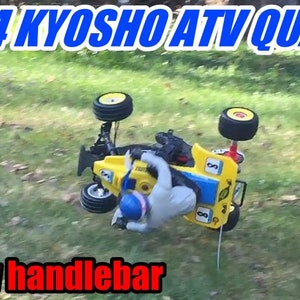 Puede incluir: Un quad ATV Kyosho de escala 1/4, de color amarillo y azul, está volcado de lado. El ATV tiene un manillar negro y ruedas negras. El ATV está sobre una superficie cubierta de hierba. El texto "New handlebar" es visible en la esquina inferior izquierda de la imagen.