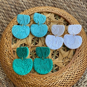 May include: A set of six earrings featuring a jungle theme. The earrings are made of polymer clay and have a two-tiered design. The top tier features a variety of jungle animals, while the bottom tier features a leafy pattern. The earrings are available in three colors: teal, green, and white.