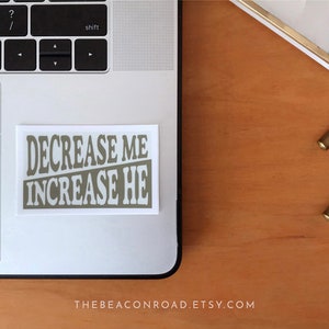 Puede incluir: Una pegatina blanca con el texto "DECREASE ME INCREASE HE" en color marr&oacute;n claro. La pegatina est&aacute; en un port&aacute;til plateado.