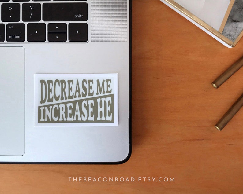 Puede incluir: Una pegatina blanca con el texto "DECREASE ME INCREASE HE" en color marr&oacute;n claro. La pegatina est&aacute; en un port&aacute;til plateado.