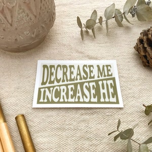 May include: A white sticker with the text "DECREASE ME INCREASE HE" in brown letters. The sticker is on a beige fabric surface.