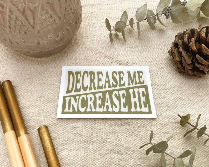 Rectangular shape with text that reads DECREASE ME INCREASE HE with inverse colors. Sticker is shown in flat lay with eucalyptus branches, pinecone, pencils, and small vase.
