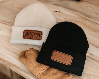 May include: Two knitted beanies, one cream and one black, with brown leather name patches. The cream beanie has "MADISON" and the black beanie has "BROOKS" printed on the patches. A brown knitted scarf is also visible.