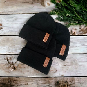 May include: Three black knit beanies with brown leather tags. One tag reads "MAMA", another "GRANDMA", and the third "ANNIE". The beanies are folded at the brim and are arranged on a wooden surface.