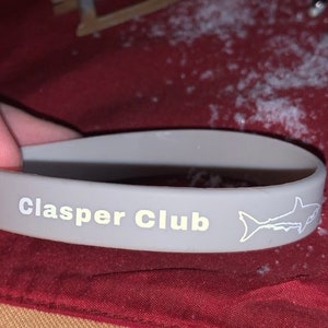 Pode incluir: Pulseira de silicone cinza com o texto "Clasper Club" e uma silhueta de tubarão branca.